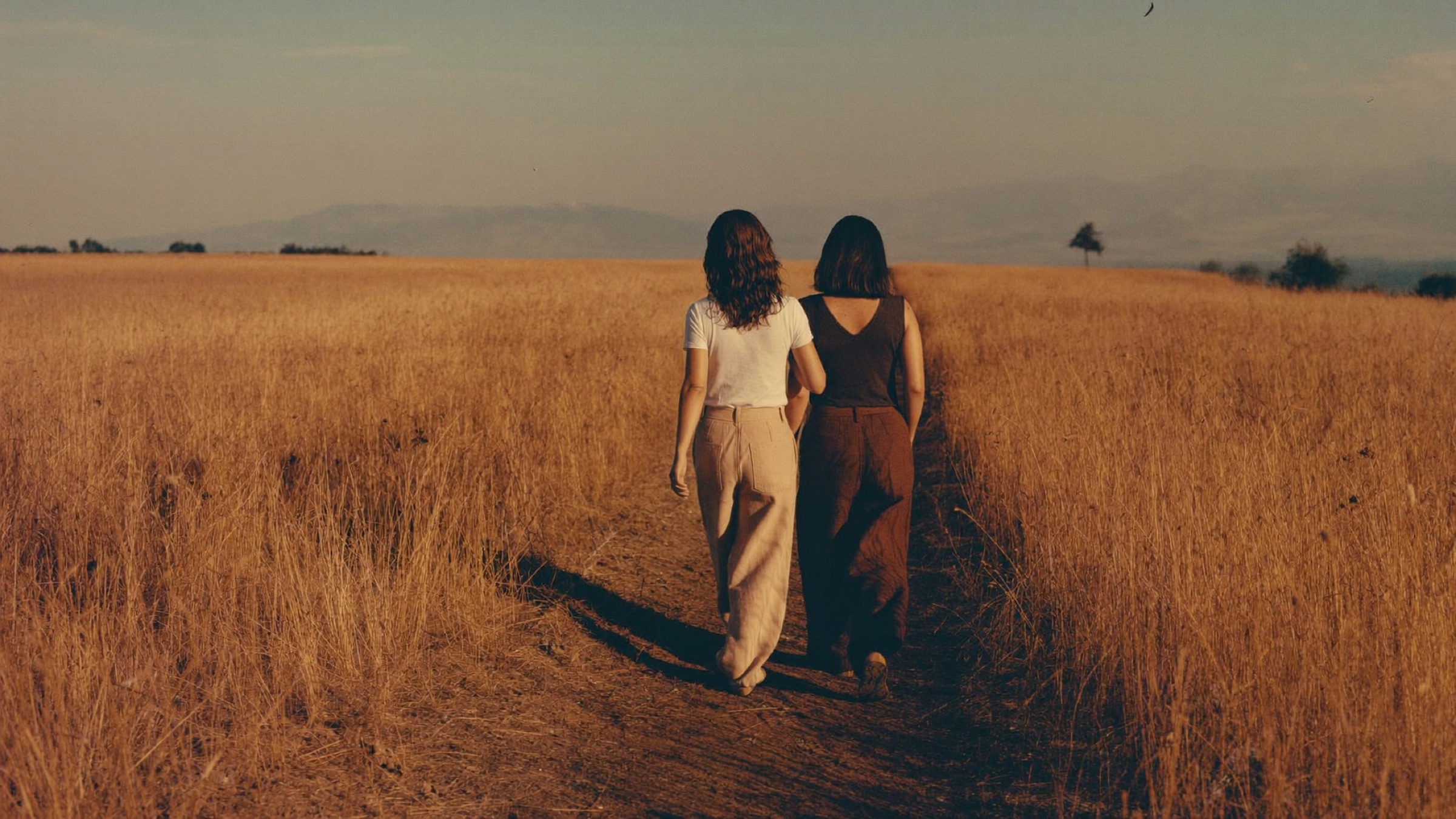 Dos mujeres caminando por una vereda de tierra entre pastos secos al atardecer
